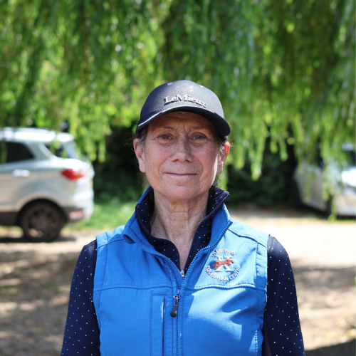 Portrait of an older woman wearing a blue vest and a black cap, standing outdoors with green trees and parked cars in the background.