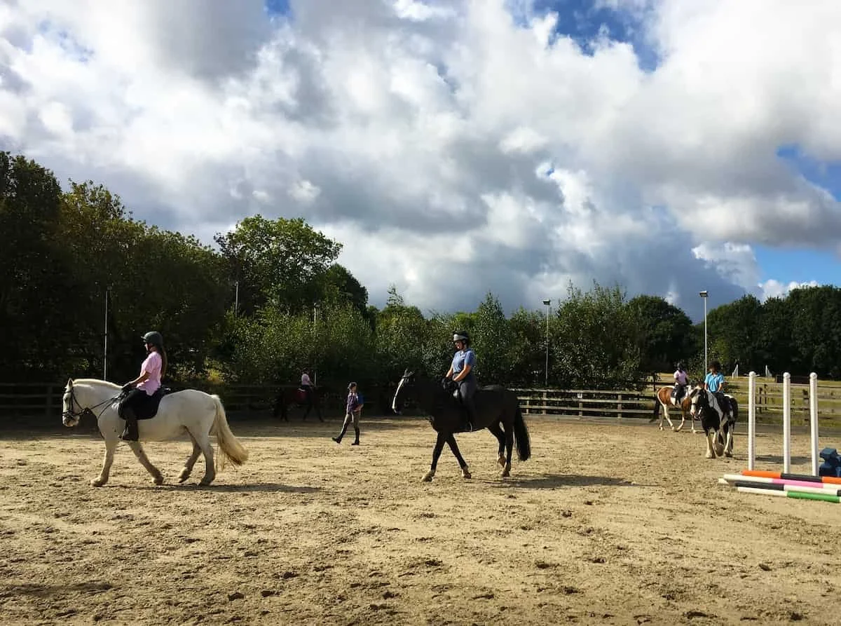 People riding horses in an outdoor riding arena with jump obstacles, trees, and cloudy sky in the background.