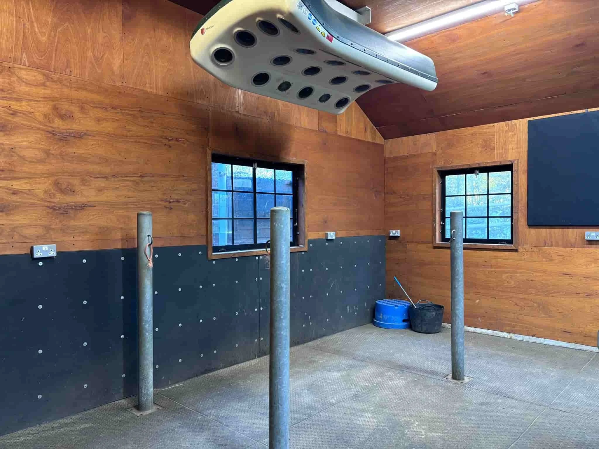 An indoor room with wooden walls and windows, empty with four metallic poles in the middle, and a bucket and a container on the floor.