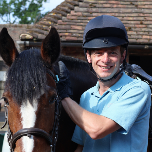A smiling man wearing a black riding helmet and gloves standing next to a brown and white horse outdoors.