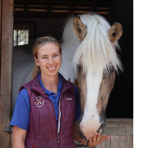 A young woman smiling next to a white horse in a stable.