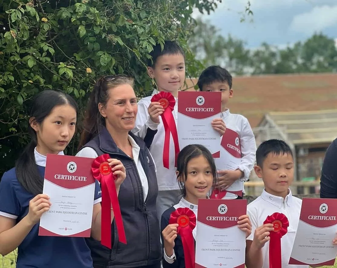 Group of children and a woman holding certificates and red ribbons outdoors, celebrating an achievement.