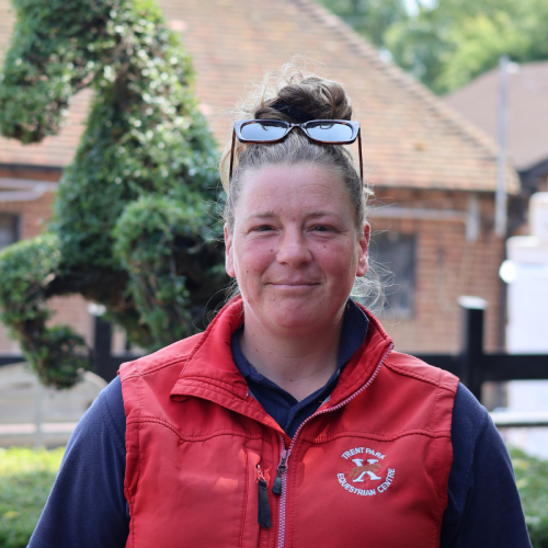 A woman outdoors wearing a red vest over a navy shirt, with sunglasses on her head, smiling with a background of trees and houses.