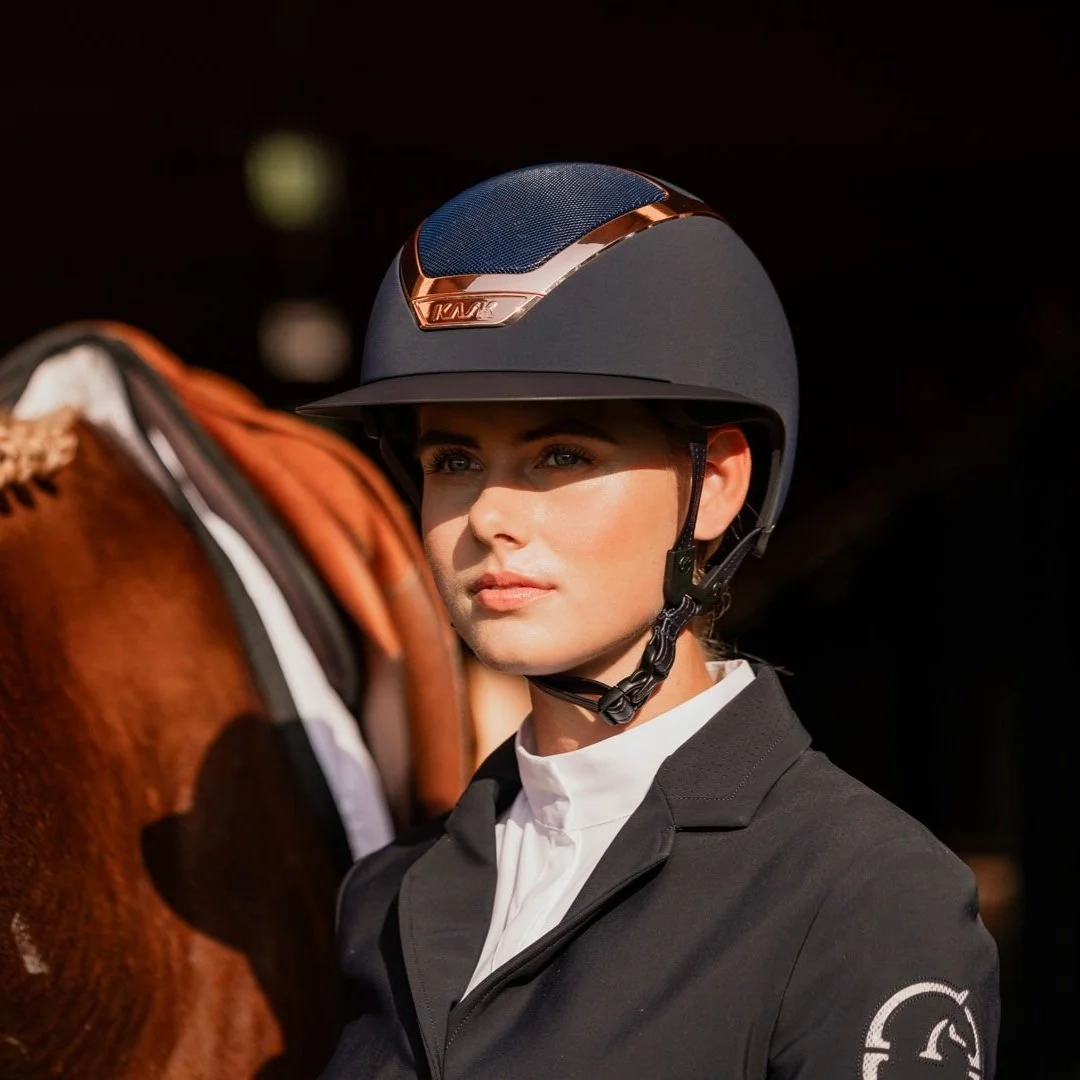 A woman wearing a black riding helmet and jacket, standing next to a brown horse.