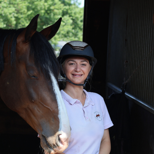 A woman in a riding helmet standing next to a brown horse inside a stable.