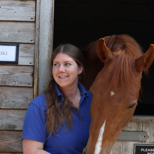 A woman with long brown hair and a blue polo shirt standing next to a brown horse outside a wooden stable.