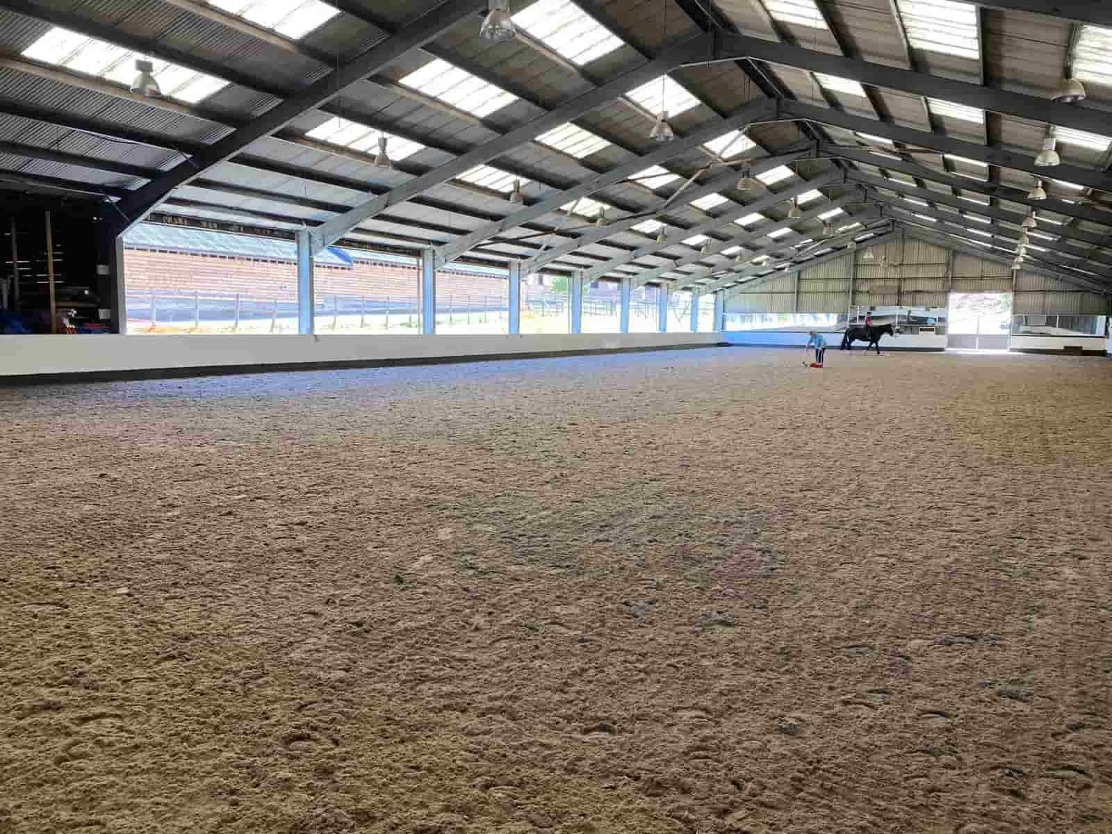An indoor horse riding arena with a sandy surface, metal roof, large windows, and a few people and a horse in the background.