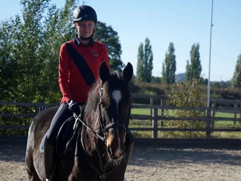 A young girl wearing a riding helmet and red jacket riding a dark horse in an outdoor riding arena.
