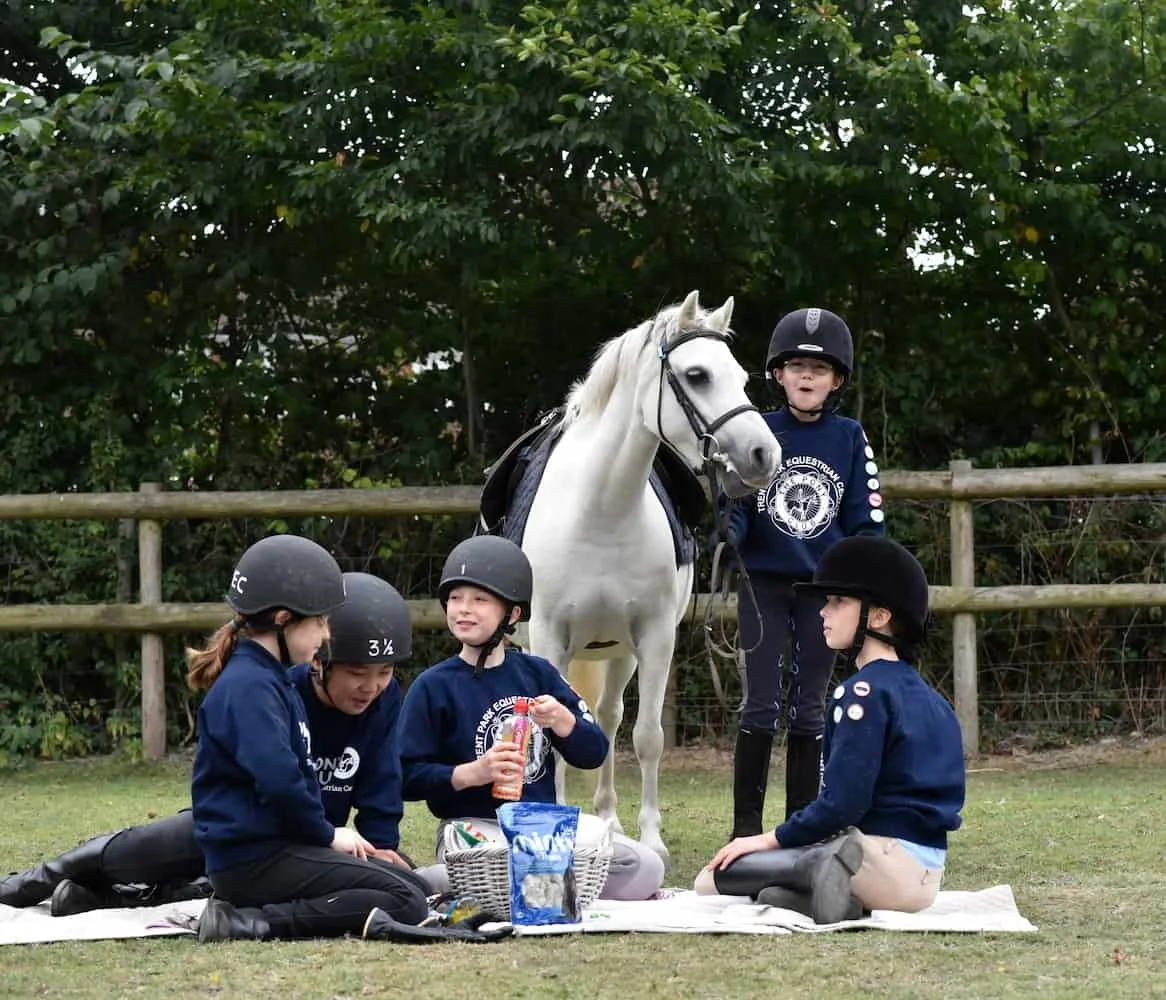 Four children in equestrian attire sitting on a blanket with a basket and snacks, and a young girl standing beside a white horse in a paddock with trees and a fence in the background.