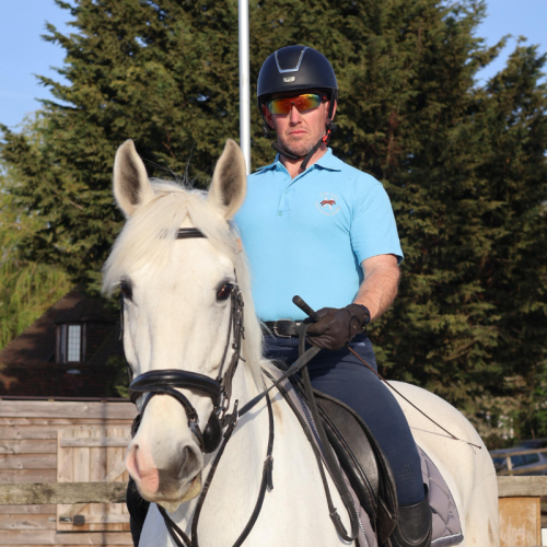 A man wearing a helmet and sunglasses riding a white horse with a black bridle outdoors.