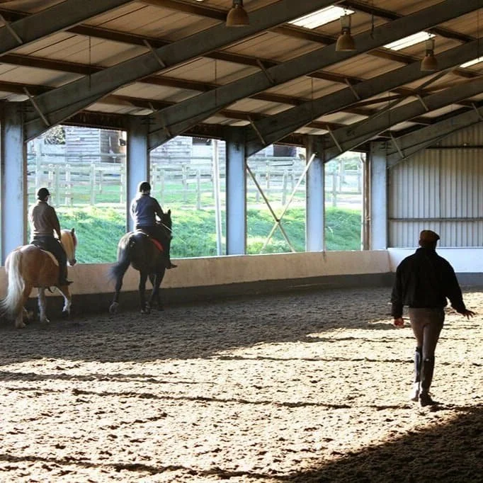 Two people riding horses inside a large indoor riding arena while a third person walks on the ground.
