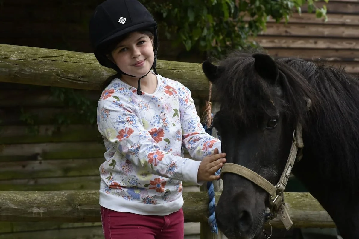 A young girl with a black riding helmet and floral long-sleeve shirt gently touching a black horse's face. They are outdoors near a wooden fence.