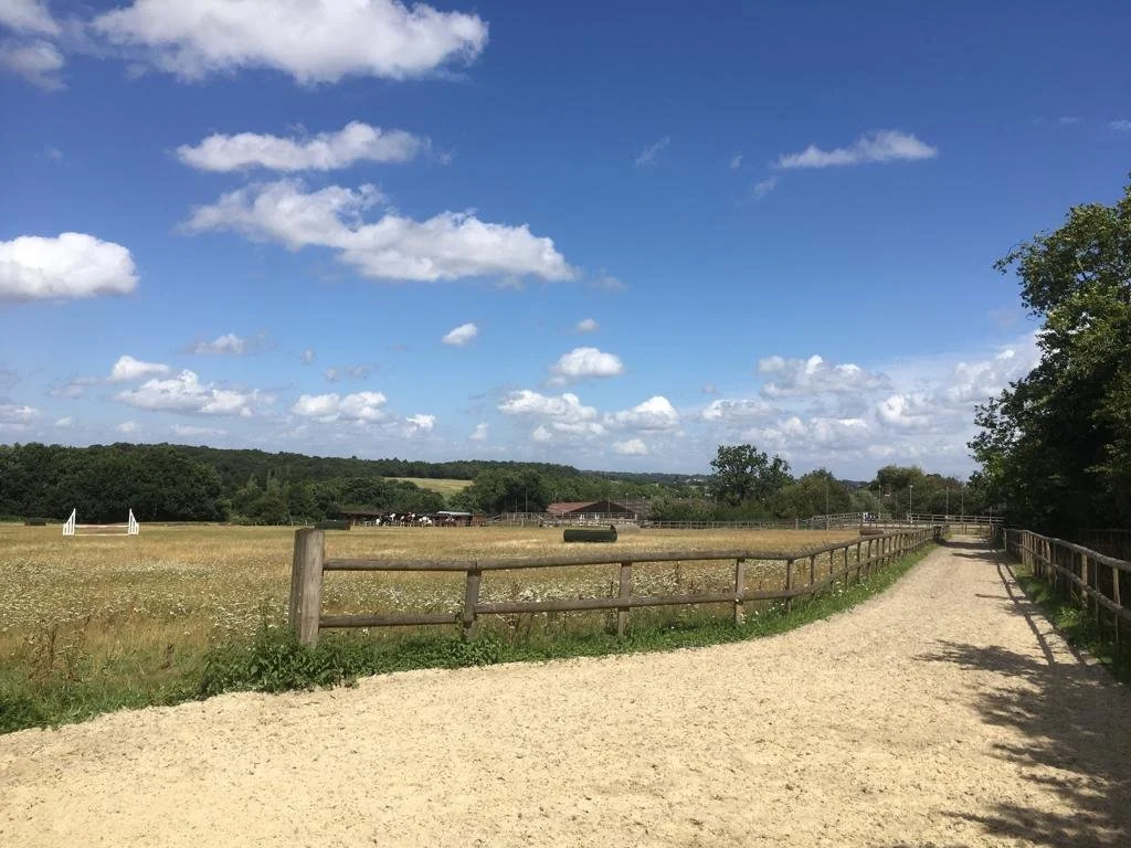 A scenic rural landscape featuring a dirt path on the right, a wooden fence lining a green pasture with white hurdles, a barn or stable in the distance, and a bright blue sky with scattered white clouds.