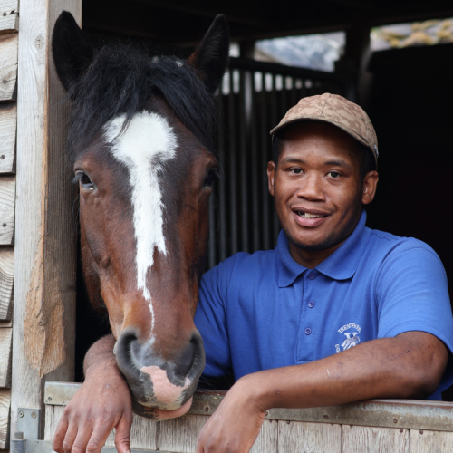 A young man in a blue polo shirt and beige cap smiling with his arm around a brown and white horse in a stable.