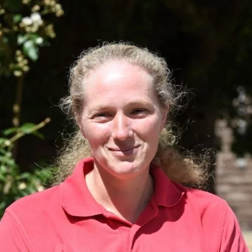 A woman with curly blonde hair wearing a red polo shirt outdoors with greenery and trees in the background.