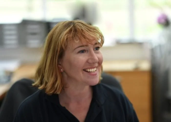 Smiling woman with short red hair, wearing a black shirt, in an office setting.