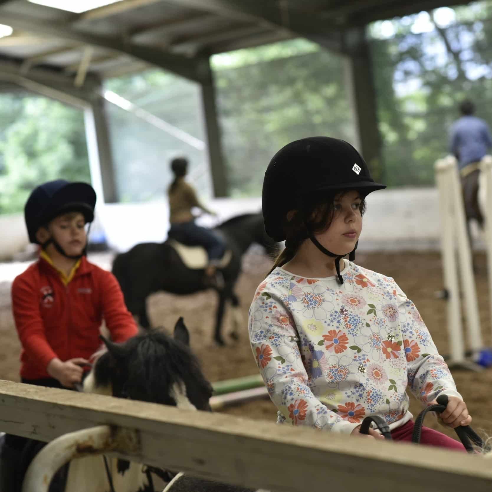 Children riding ponies at an indoor riding arena, with one girl in the foreground wearing a black riding helmet and floral shirt, and other children and ponies in the background.