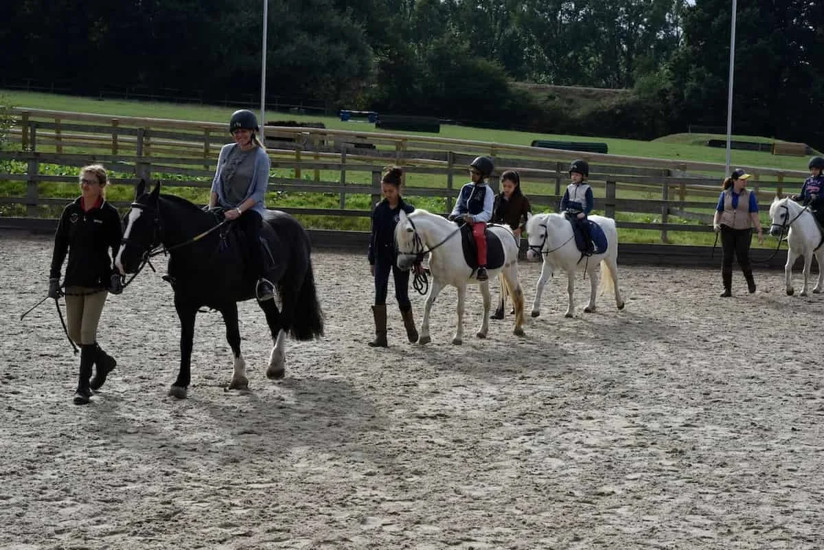 A group of people riding horses in an outdoor riding arena, with wooden fences and trees in the background.