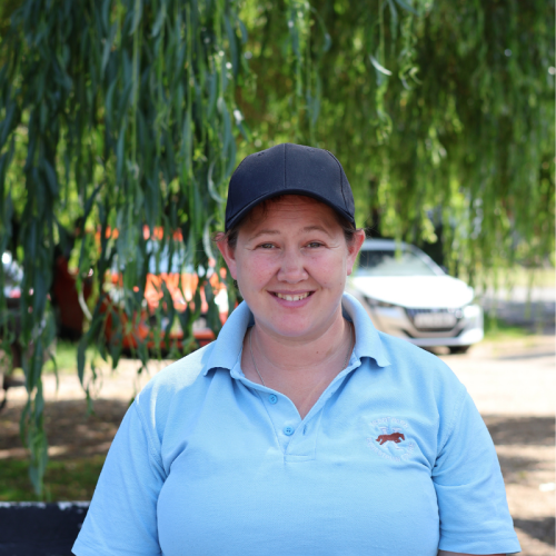 A woman smiling outdoors, wearing a navy baseball cap and a light blue polo shirt, standing under a leafy tree with a blurred background of a car and trees.