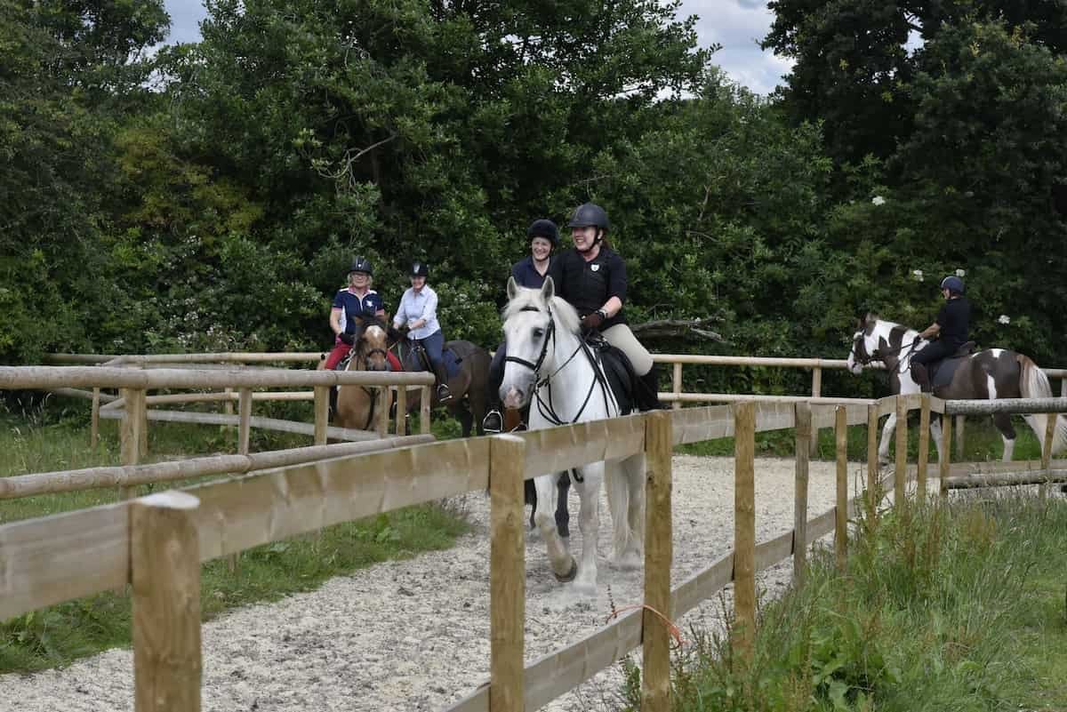 Four people riding horses in a fenced outdoor riding arena with trees in the background, two of whom are smiling at the camera.