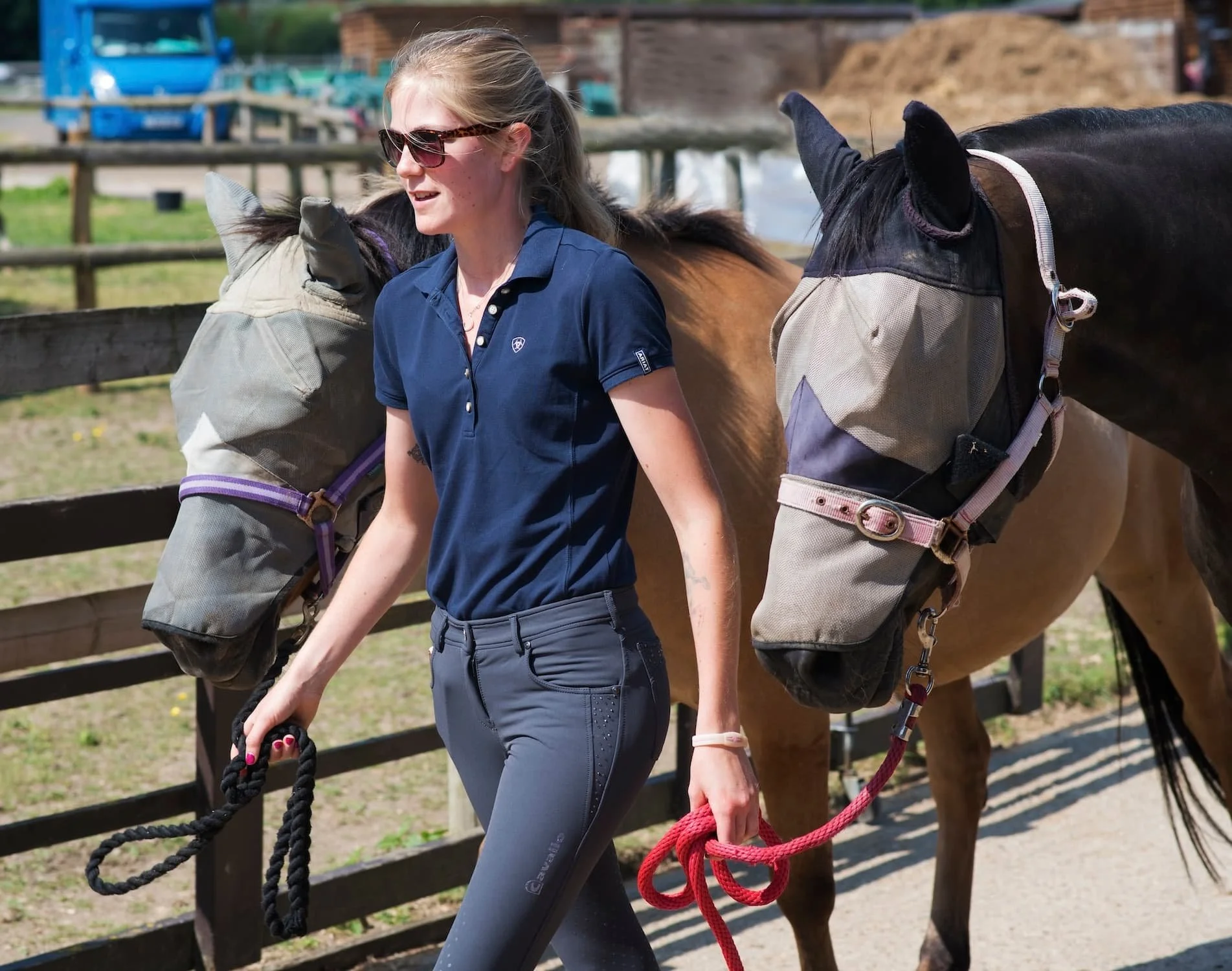 A woman with blonde hair, sunglasses, and a navy polo shirt leading a brown horse with a black mane, both wearing halters, inside a fenced outdoor area with wooden structures and a blue truck in the background.