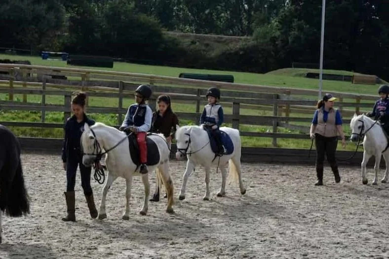 Children riding white horses in a fenced outdoor riding arena, with instructors guiding them.