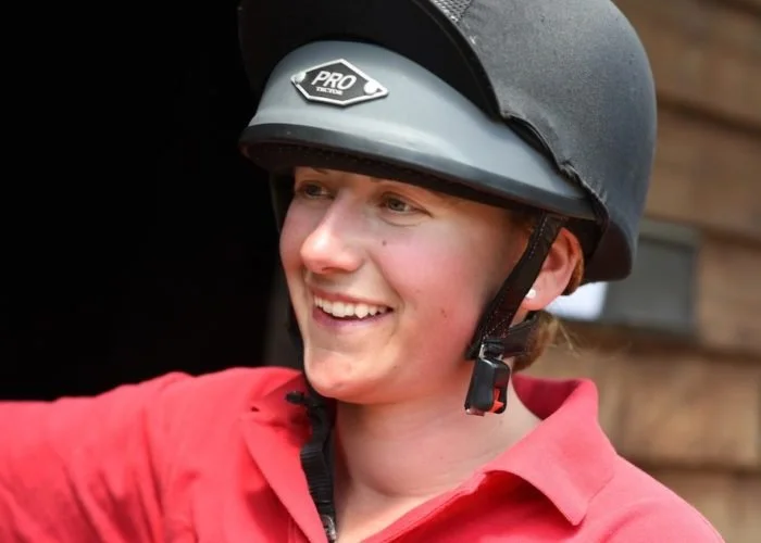 A young woman smiling and wearing a black helmet with a gray sticker labeled "PRO" in front of a wooden wall.