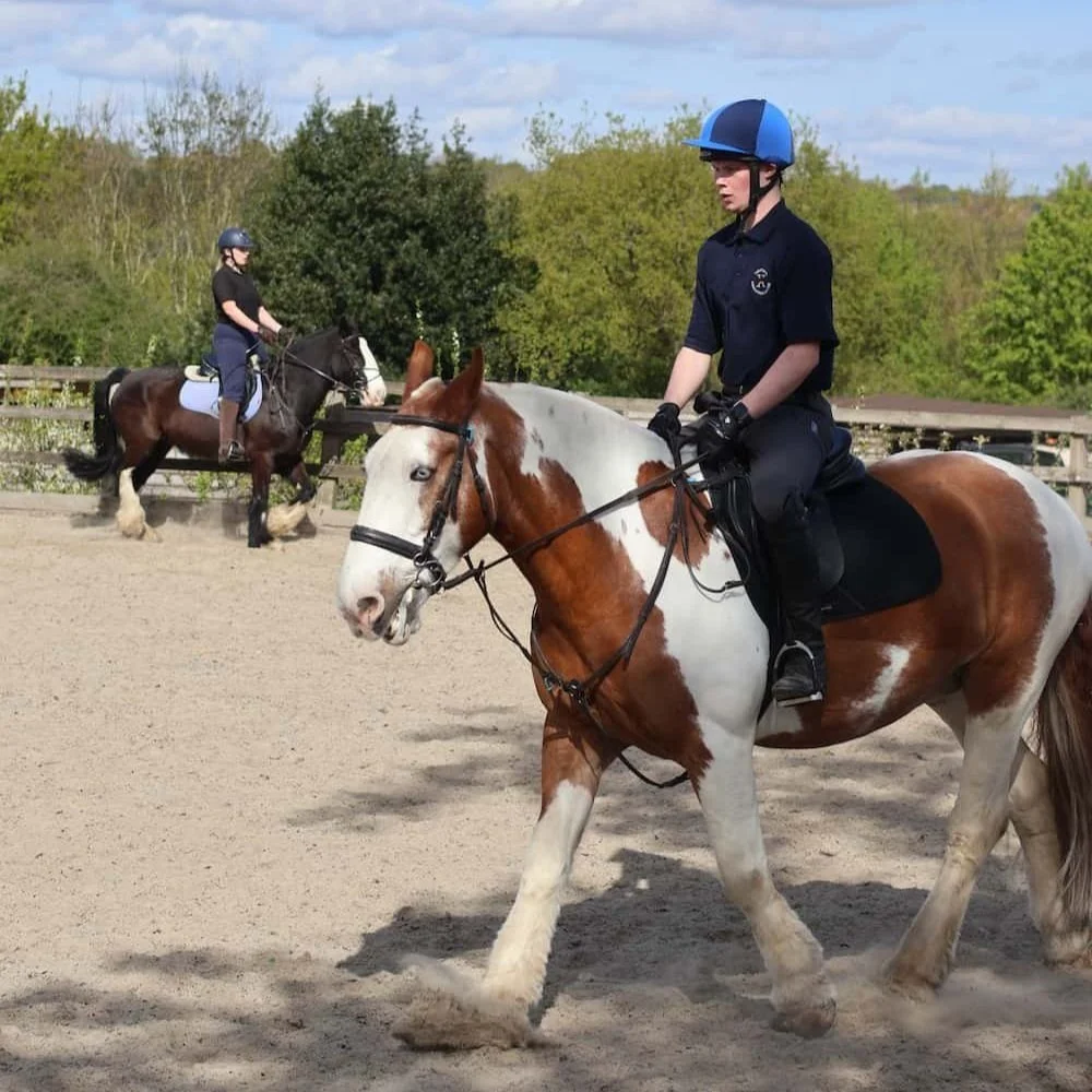 A young man riding a paint horse with brown and white patches during a horseback riding lesson or practice, with a woman riding a dark brown horse in the background at an outdoor riding arena.