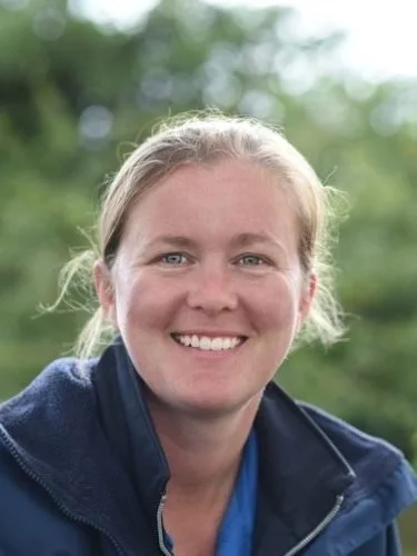 Smiling woman with blonde hair and blue eyes outdoors, wearing a navy jacket.