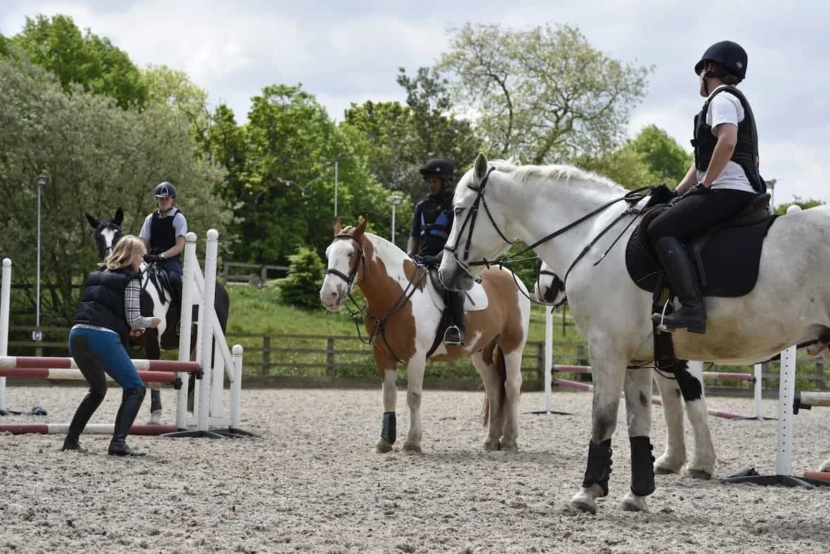People riding horses in an outdoor equestrian training or riding area, with an instructor guiding a rider during a lesson.