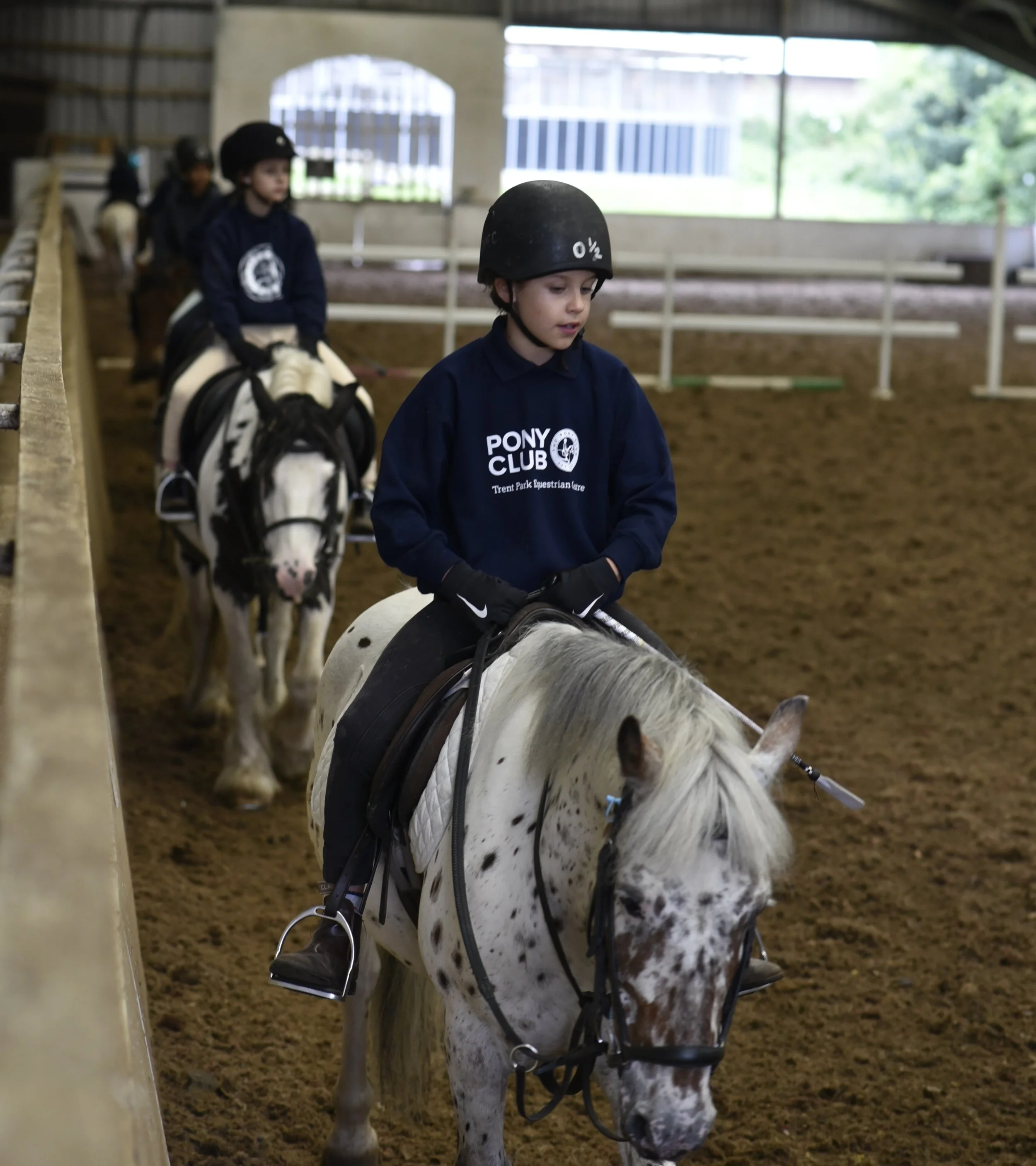 Children riding ponies in an indoor riding arena. The boy in front wears a black helmet and a navy hoodie that reads 'Pony Club,' with other children visible in the background.