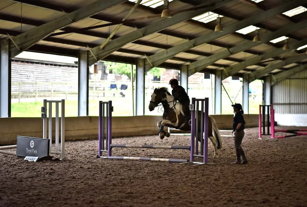 A person riding a horse jumps over an obstacle in an indoor horse riding arena next to a trainer or instructor.