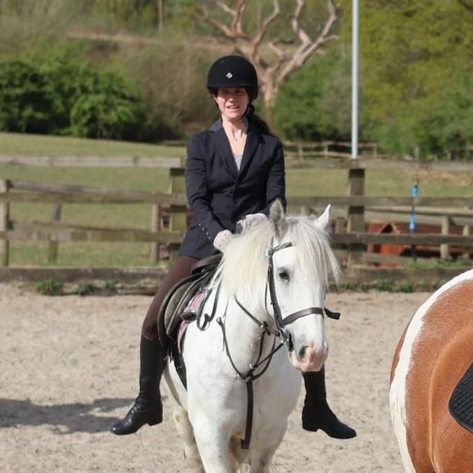 A woman in riding gear, including a helmet, smiles while riding a white horse in an outdoor riding arena.