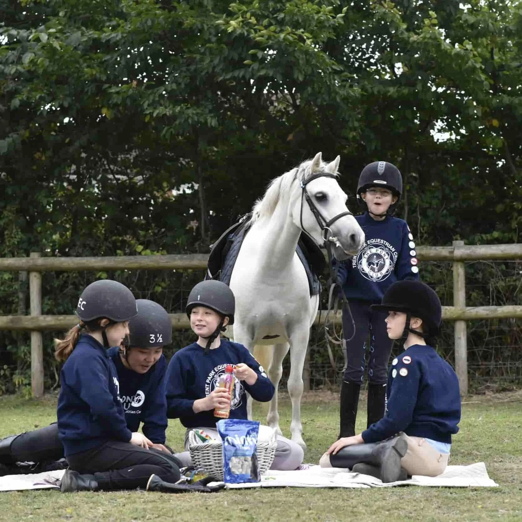 A group of children in navy blue jackets wearing black helmets, sitting and standing on grass near a wooden fence, with a white horse behind them. The children are outdoors with green trees in the background, and there are snacks and a picnic basket on a blanket on the ground.