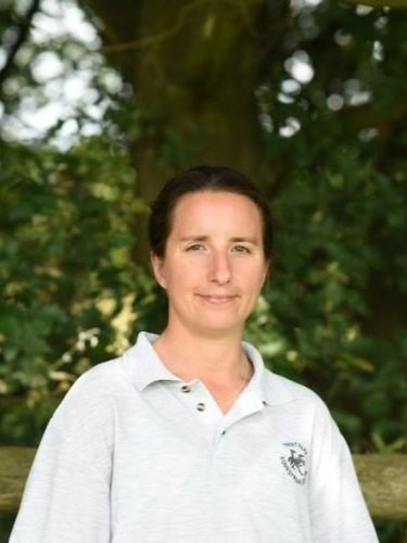 A woman with brown hair pulled back, wearing a white collared shirt, standing outdoors in front of green foliage and trees.