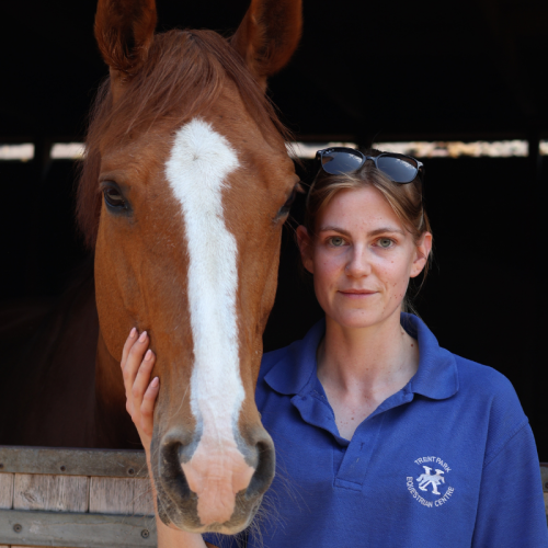 A woman in a blue shirt with sunglasses on her head standing next to a horse with a white stripe on its face inside a barn.