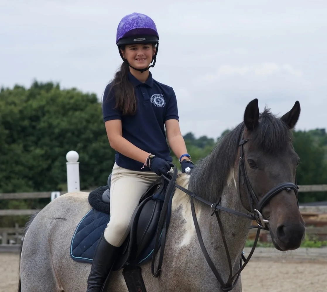 A girl riding a gray horse in an outdoor riding arena, wearing a purple helmet, navy polo shirt, beige riding pants, and black riding boots.