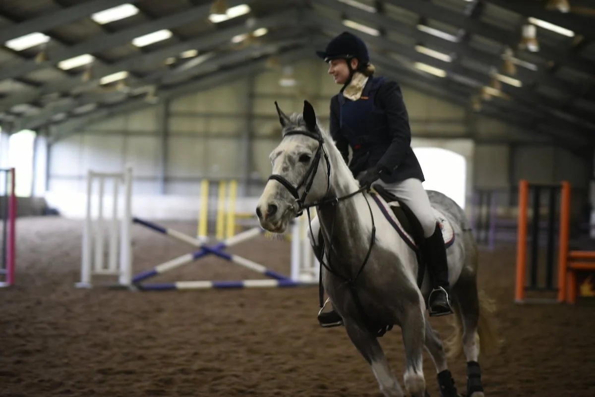 A woman riding a gray horse inside an indoor equestrian arena. She is wearing a helmet, a dark jacket, and white pants, smiling as she rides.