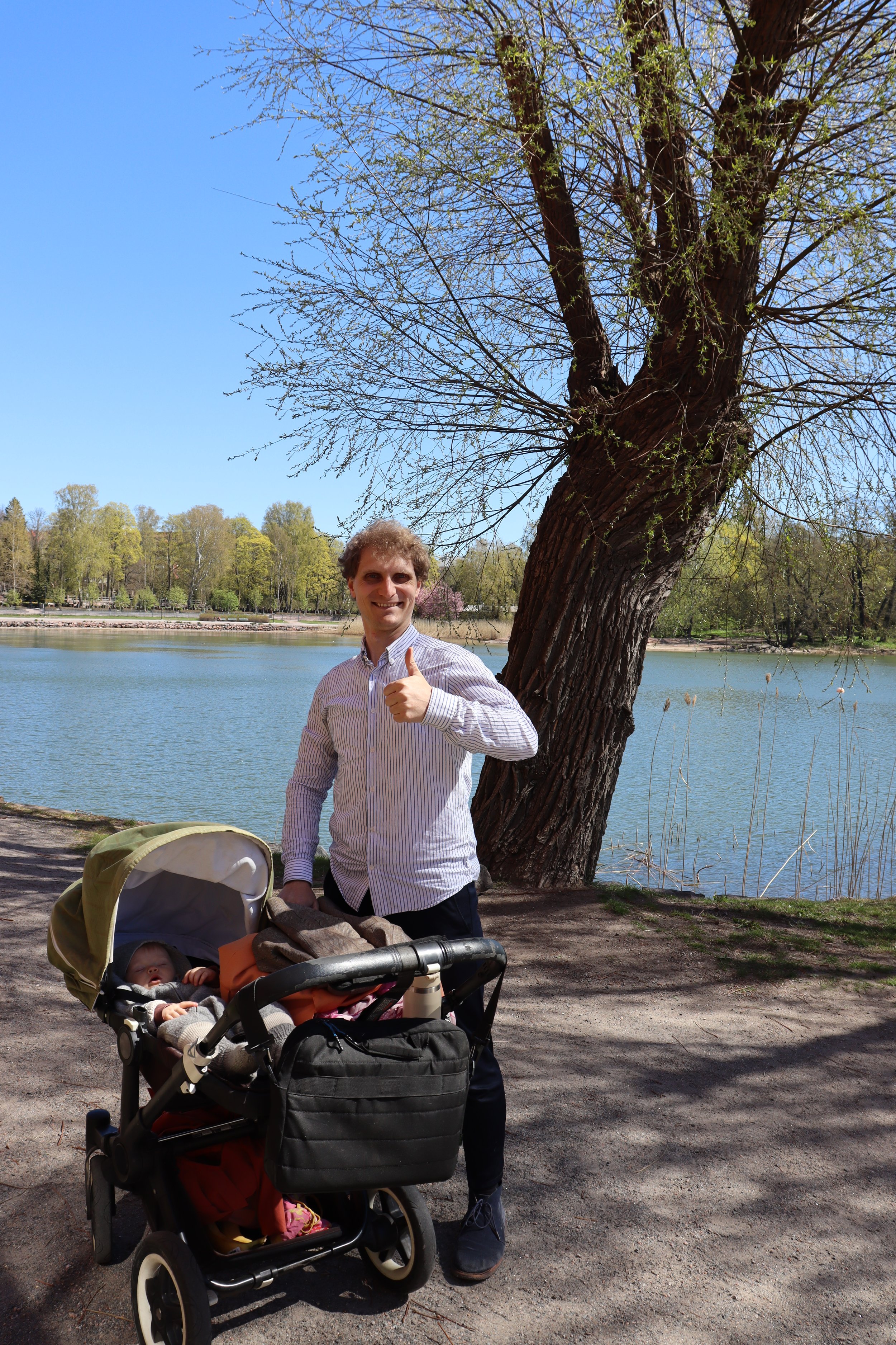 Mies seisoo puiston reunassa, hymyilee ja pitelee kädessään peukalon ylös. Hänellä on vaaleanraidallinen paita ja tummat housut. Hänellä on rattaissa nukkuva vauva ja taustalla meri, puhdas vesi ja vihreitä puita, sekä sininen taivas.