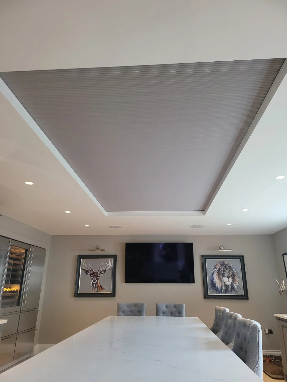 Modern dining room with a large white marble table and gray upholstered chairs. The walls feature framed animal portraits, including a deer and a lion. A large flat-screen TV is mounted on the wall. The ceiling has recessed lighting and a central decorative panel.