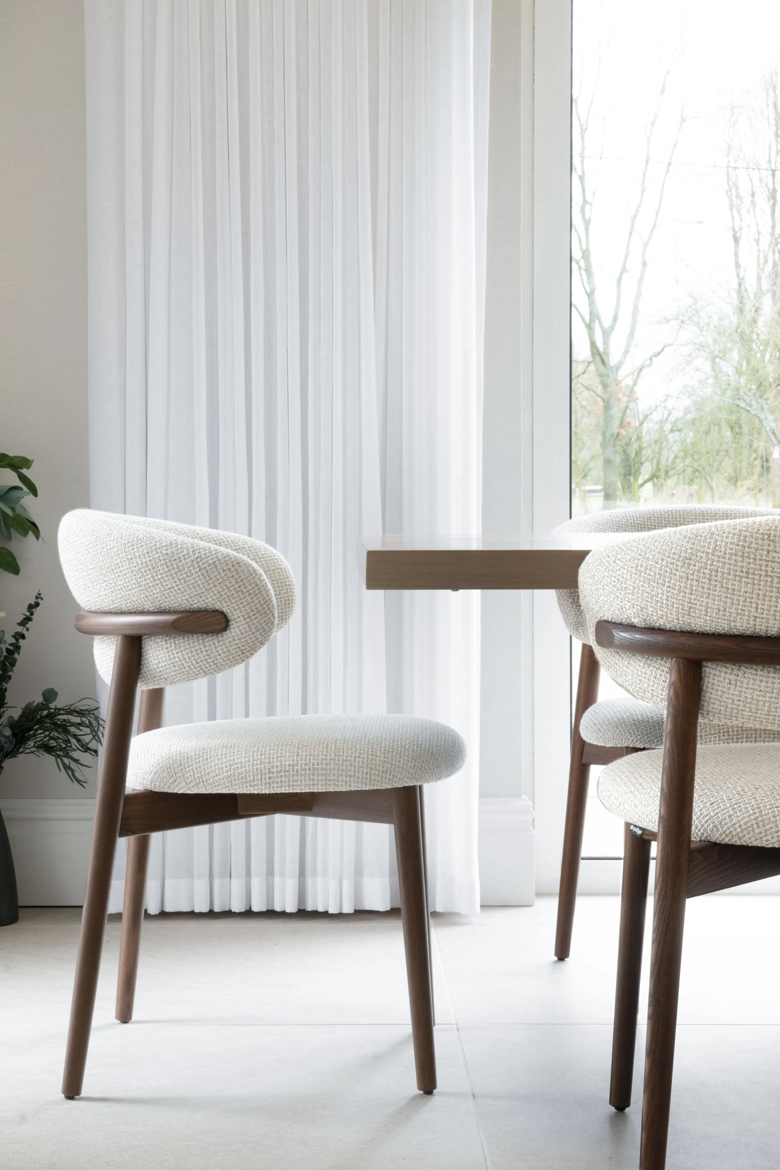 A bright room with a round table and two upholstered chairs with wooden legs, near a large window with white curtains and a view of trees outside.