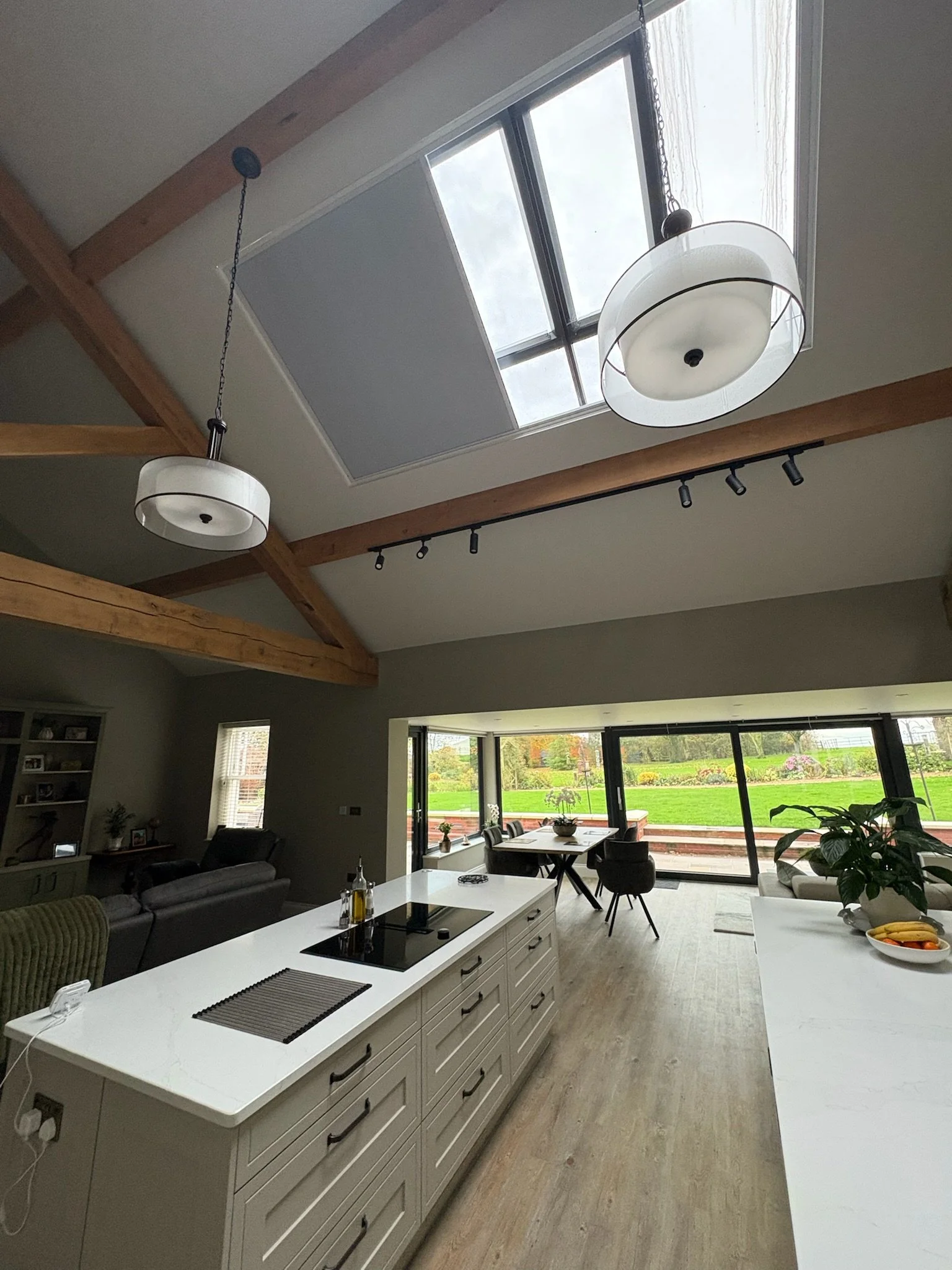 Kitchen with a white island, black cooktop, and modern lighting, overlooking a living area with large sliding glass doors that reveal a green outdoor landscape.