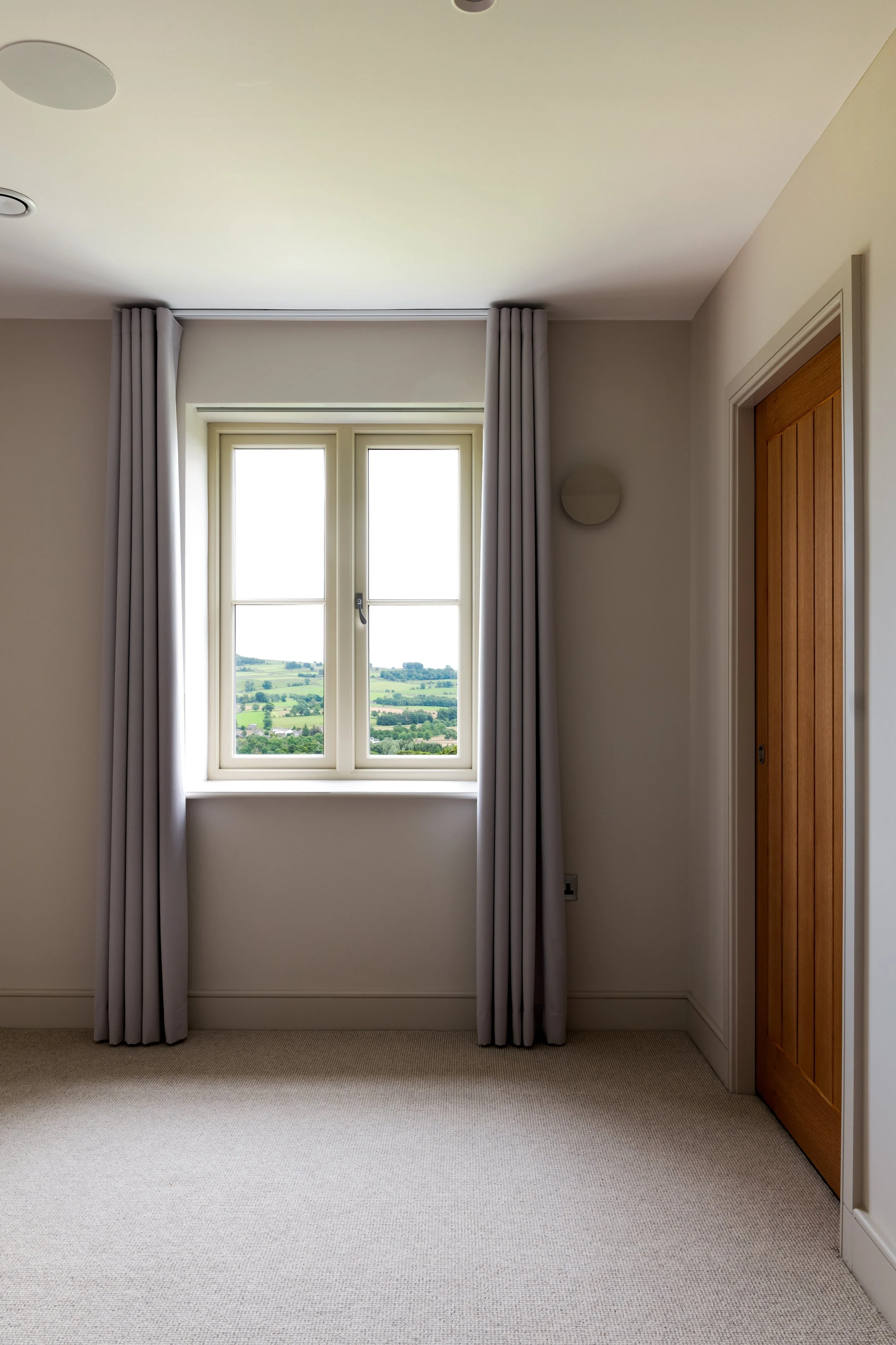 Empty room with a window showing a green landscape, beige carpeting, and gray curtains.