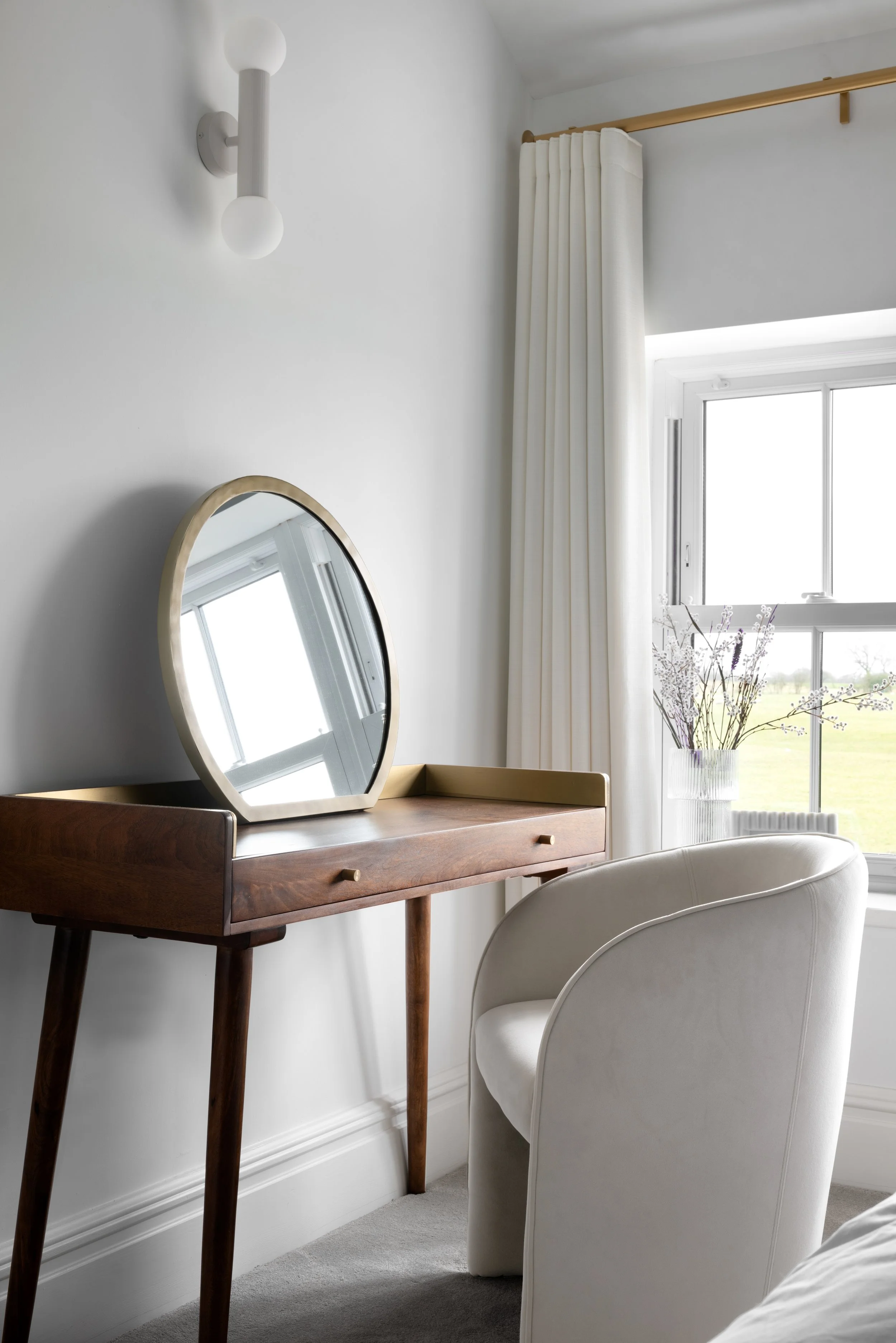 A wooden vanity table with a mirror, a white upholstered armchair, and a window with white curtains and a vase of purple flowers.