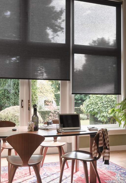 Bright home office with wooden chairs and a table, equipped with a laptop and other items, large windows with gray roller shades, view of garden outside.