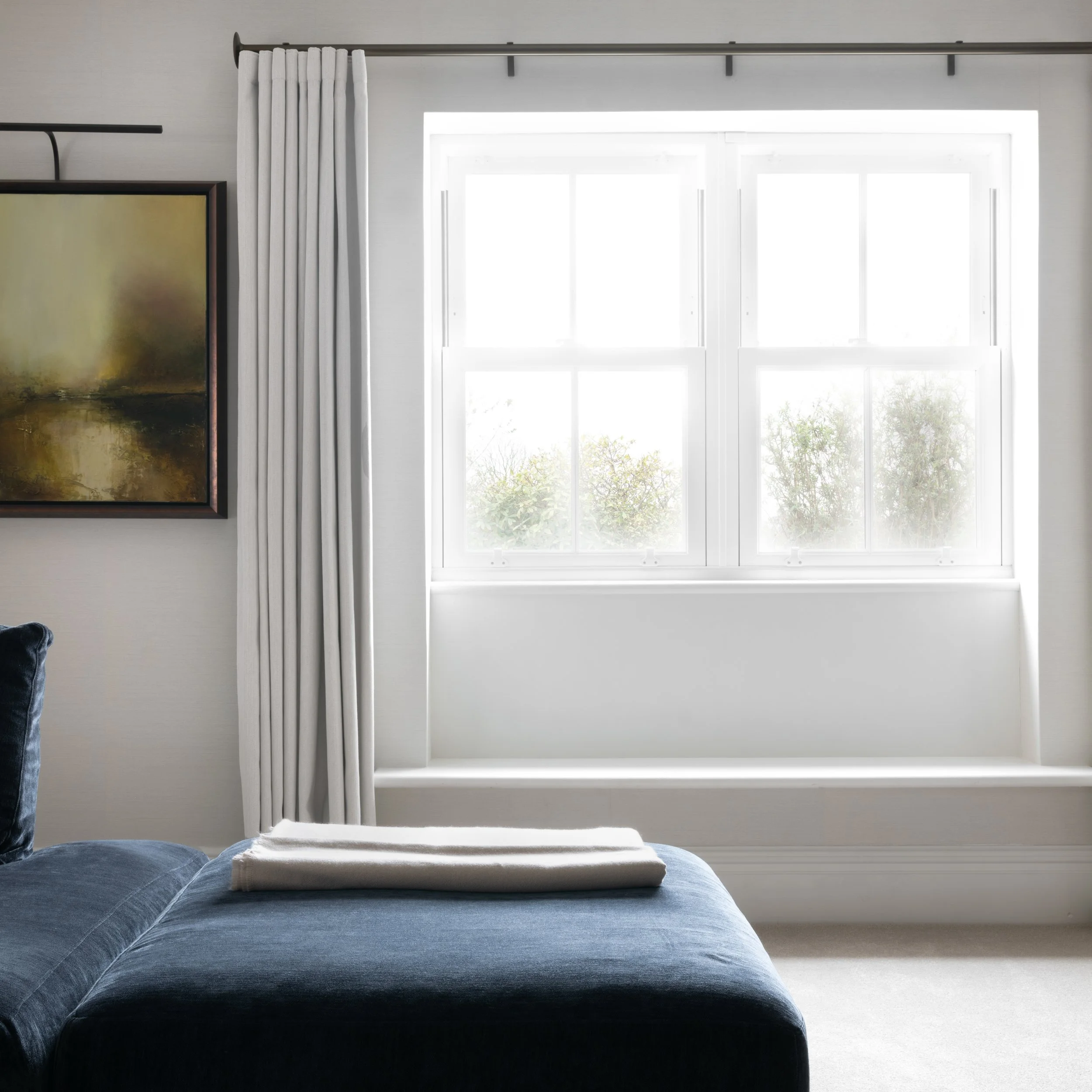 A bright living room with a large window letting in natural light, a beige bench under the window, a dark blue sofa with a blanket on it, a white curtain on the left, and a framed abstract painting on the wall.