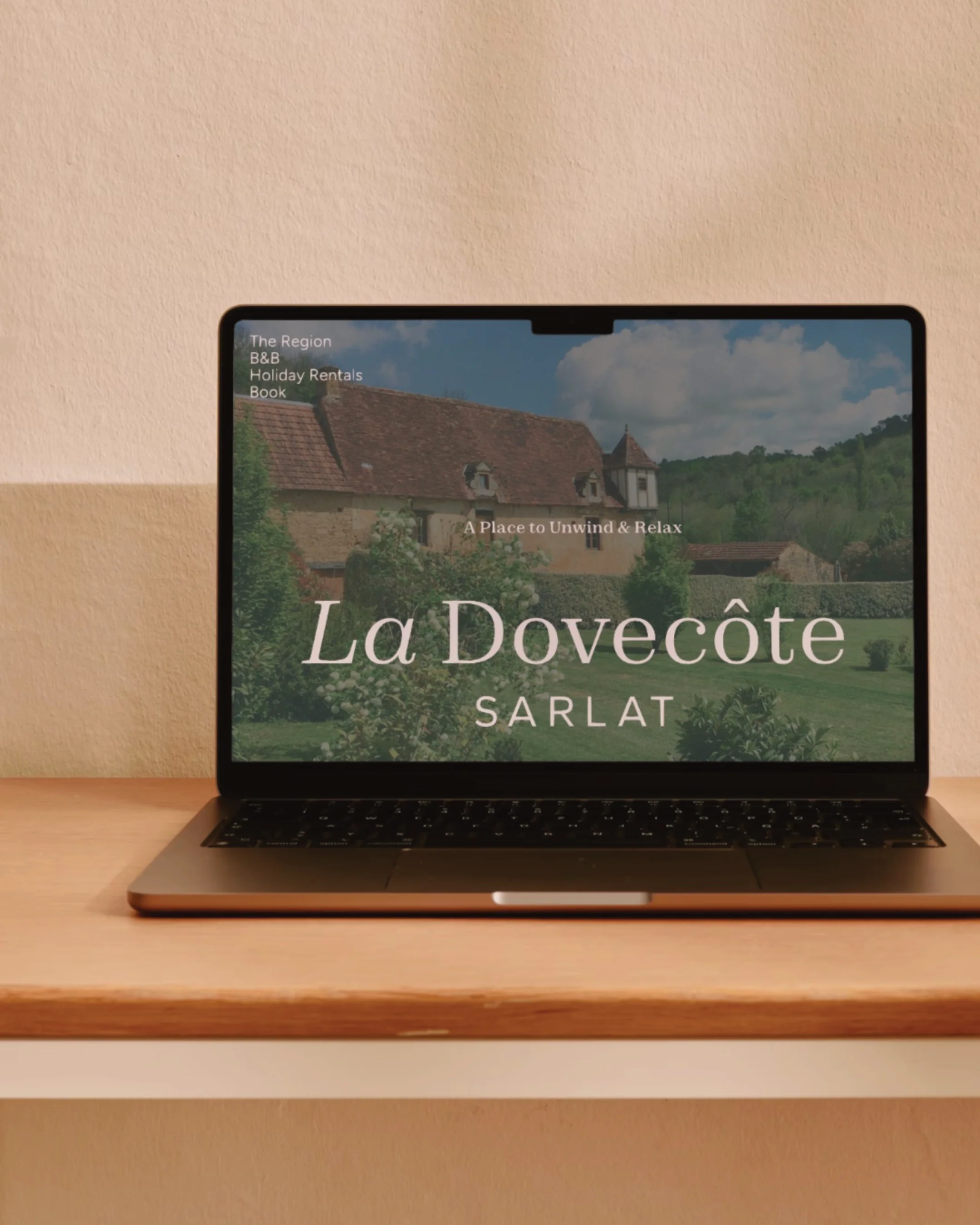 Laptop on a wooden desk displaying a website for La Dovecôte Sarlat with a background image of a historic house and garden.