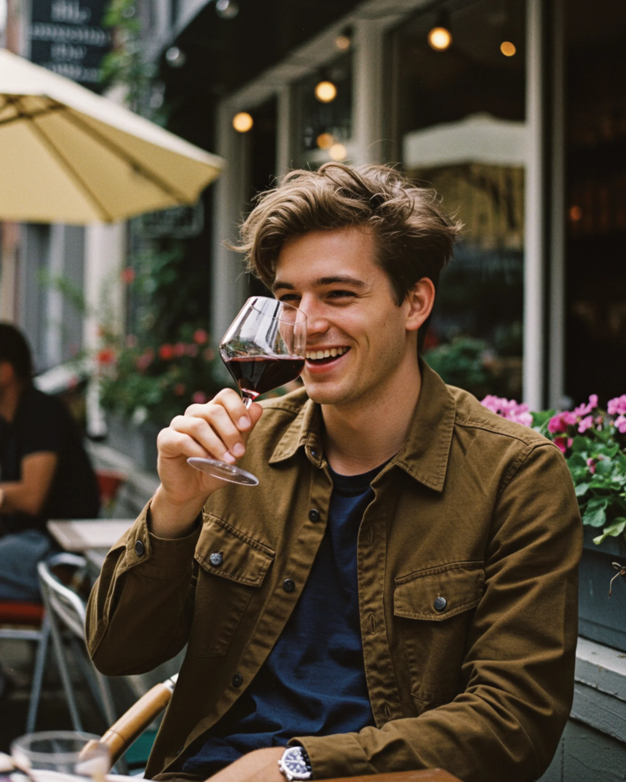 A young man smiling while holding a glass of red wine at an outdoor café with flowers and umbrellas.