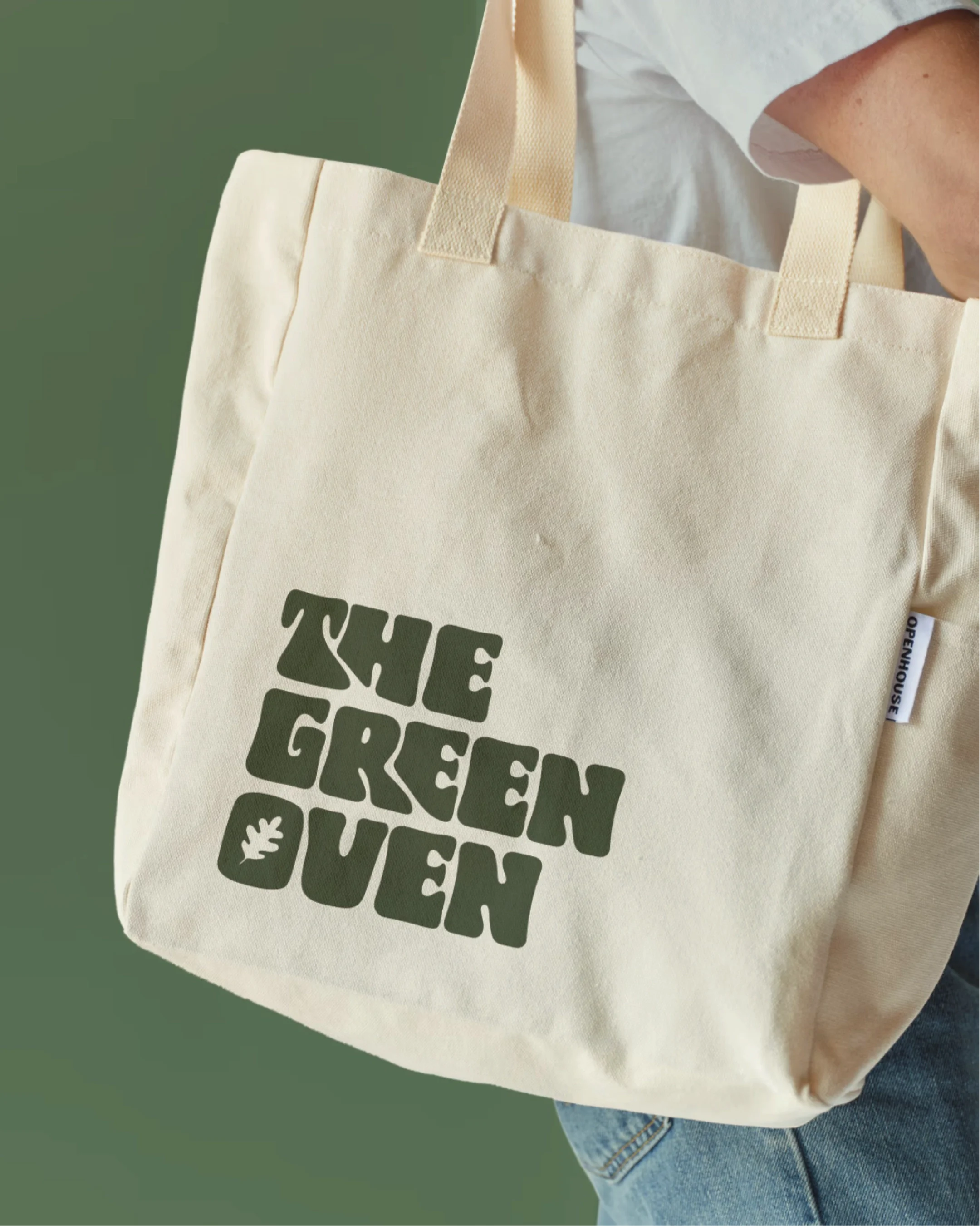 A beige tote bag with 'THE GREEN QUEEN' printed on it in green font, held by a person wearing a white shirt and blue jeans, against a green background.