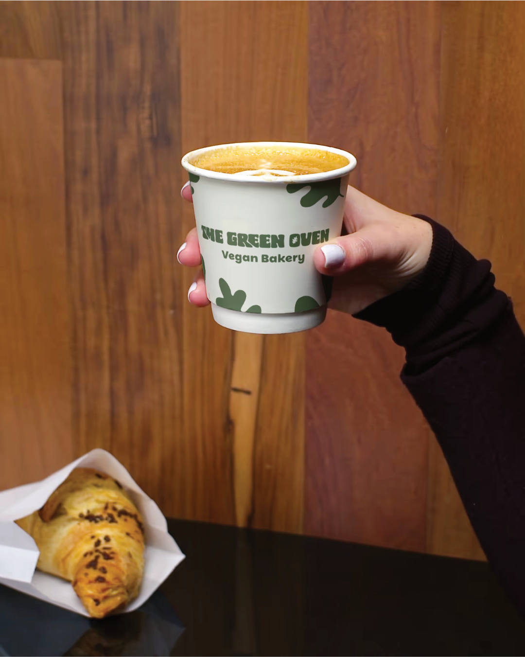 a hand holding a disposable branded coffee cup for The Green Oven, wooden wall background and a croissant on the table